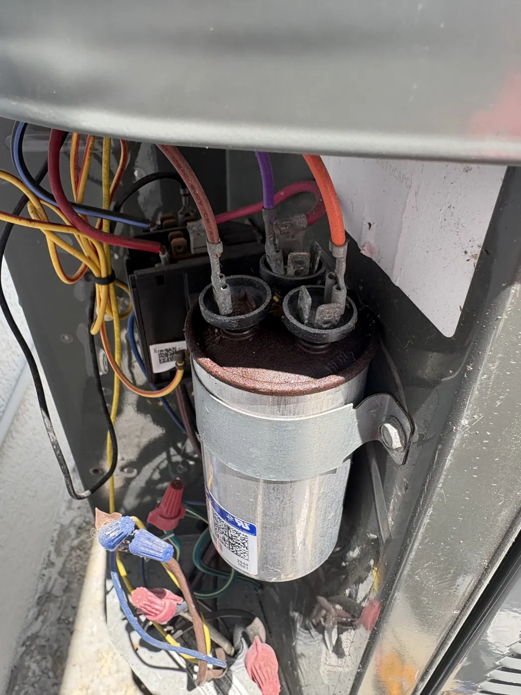 An aged dual run capacitor with a corroded top still wired in place inside a condenser electrical compartment