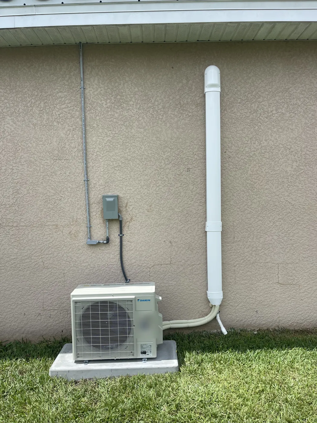 A finished ductless mini-split outdoor unit on a pad beside a stucco wall, its covered line set running up toward the soffit