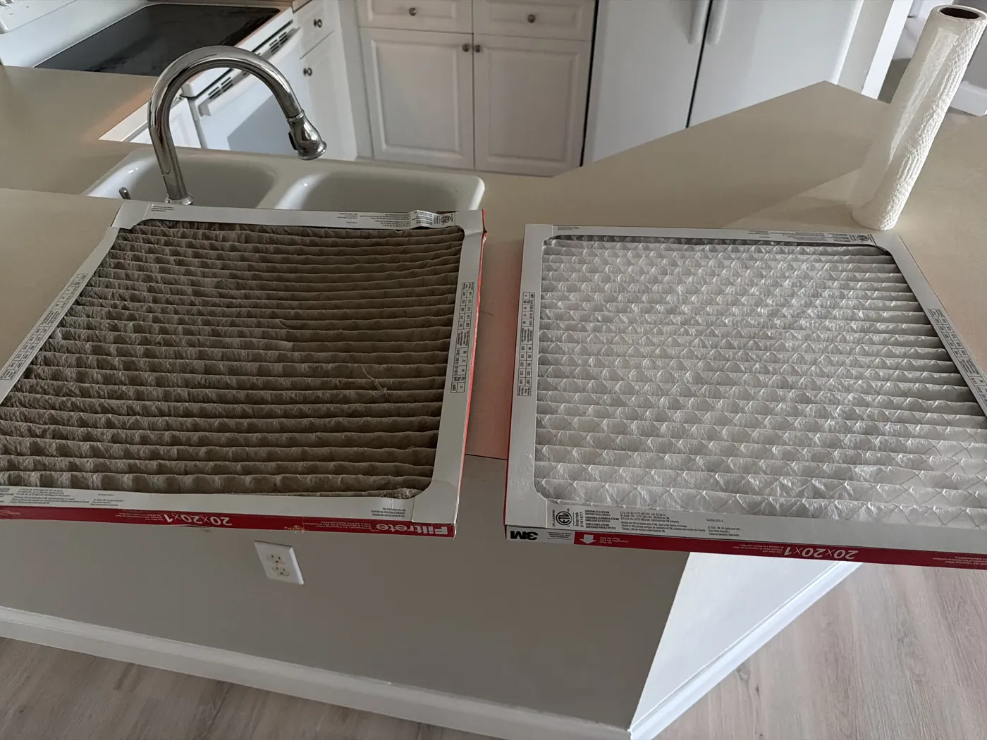 A used pleated air filter dark with dust lying next to a new white one of the same 20x20x1 size